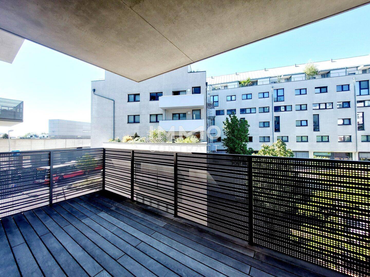 Geräumiger Balkon mit Holzbelag und Blick auf moderne Gebäude und grüne Umgebung.