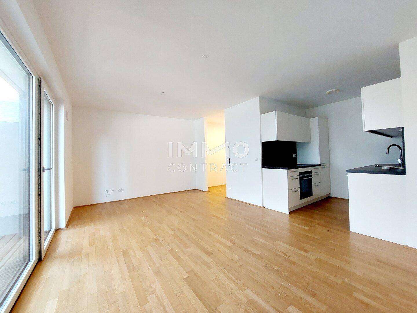 Offener Wohnbereich mit integrierter Küche, Holzboden und großen Fenstern zum Außenbereich.