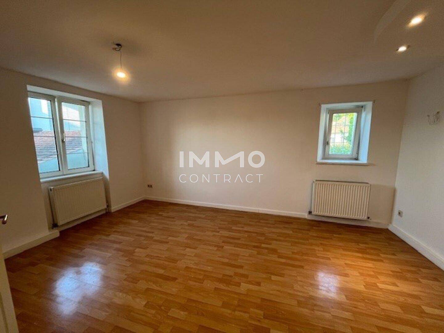 Geräumiges Wohnzimmer mit Holzparkettboden, hellen Wänden und zwei Fenstern.