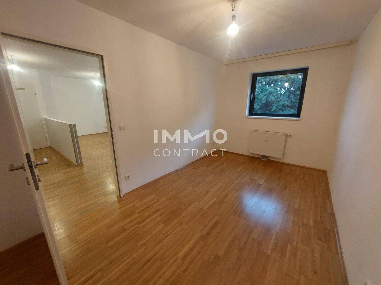 Helles Schlafzimmer mit Parkettboden und einem Fenster mit Blick ins Grüne.