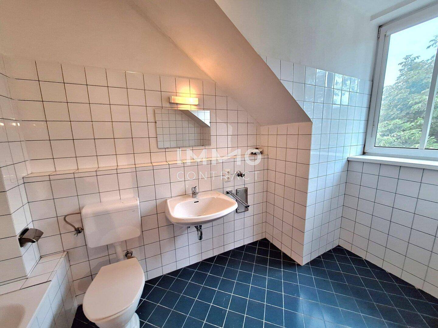 Badezimmer mit Dachschräge, WC, Waschbecken, Fenster, weißen Wandfliesen und blauen Bodenfliesen.