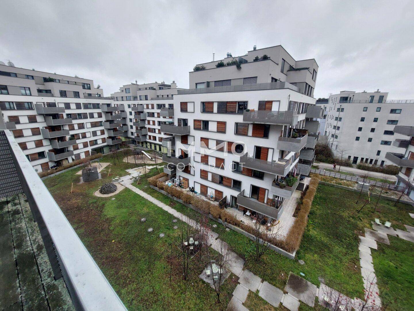 Moderne Wohnanlage mit mehreren Gebäuden, begrünten Innenhöfen und Kinderspielplatz unter bewölktem Himmel.