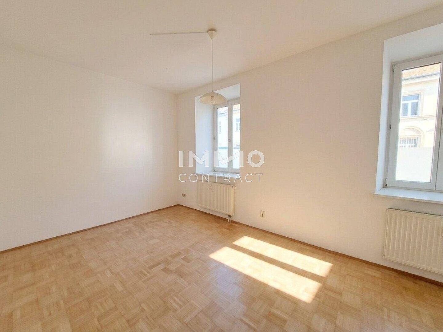 Heller Raum mit Fischgrätparkettboden, weißen Wänden und zwei Fenstern, die viel Tageslicht hereinlassen.