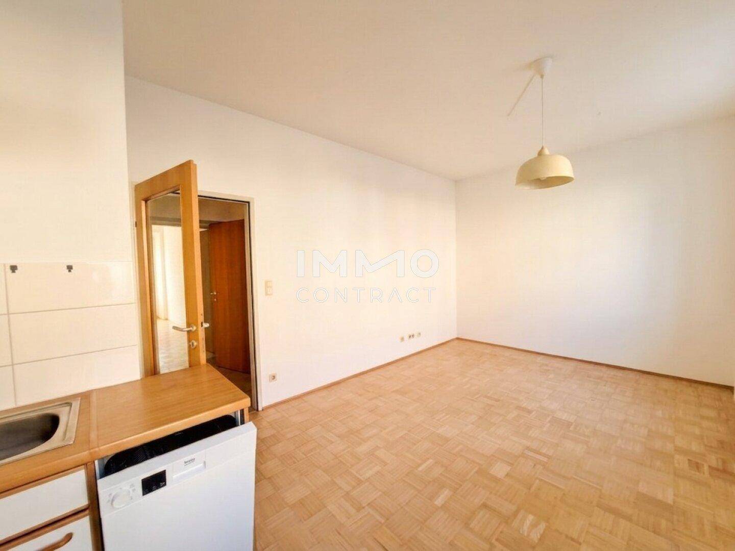 Heller Raum mit Parkettboden und einer kleinen Küchenzeile, die in den Wohnbereich integriert ist.