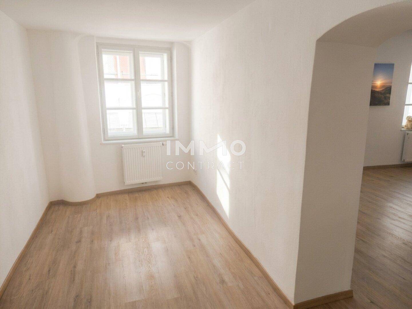 Leerer Raum mit Fenster, Heizkörper und Laminatboden, verbunden durch einen Bogen.
