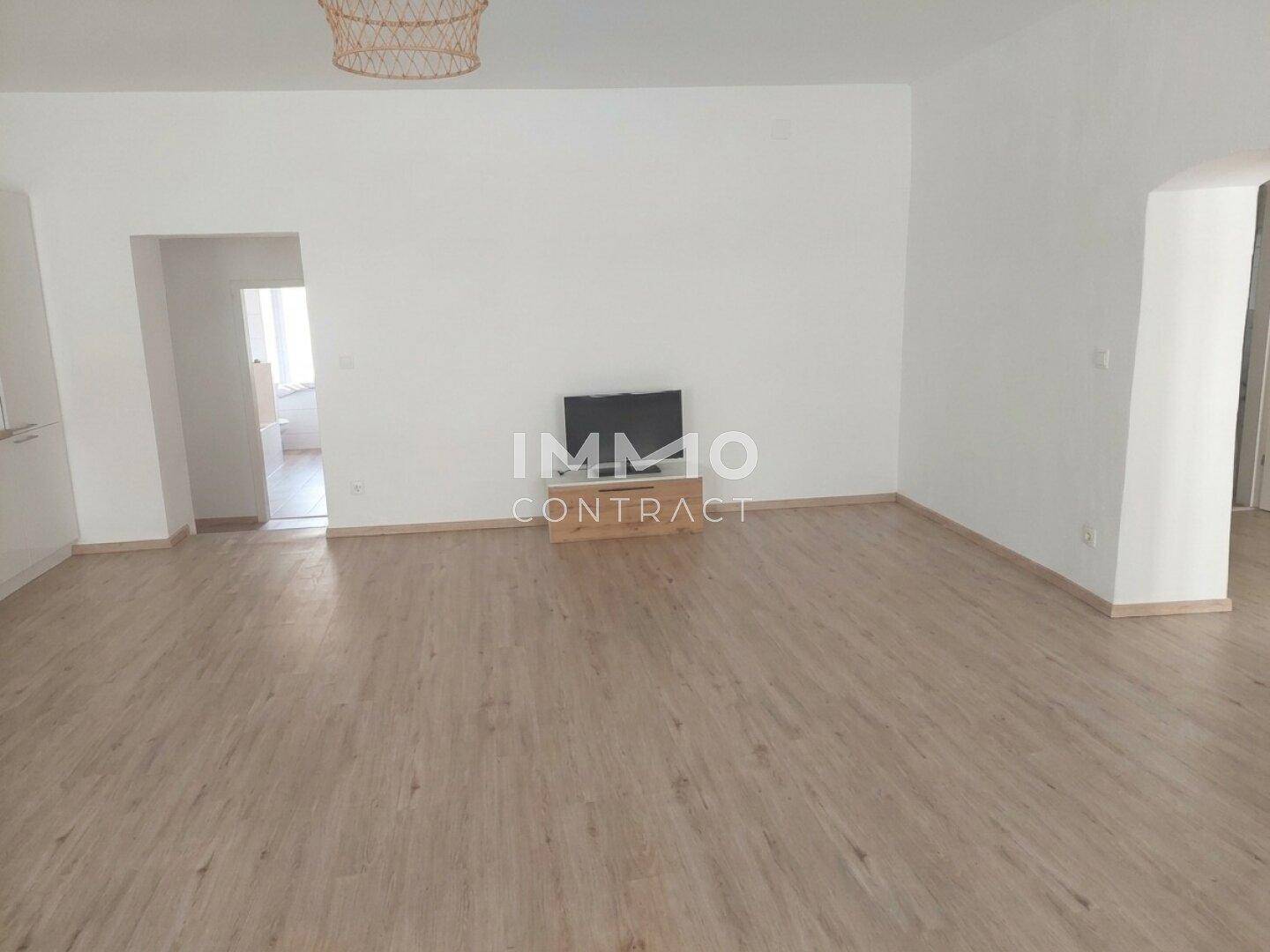 Geräumiges Wohnzimmer mit Laminatboden und Fernseher, offen zu einem weiteren Bereich.