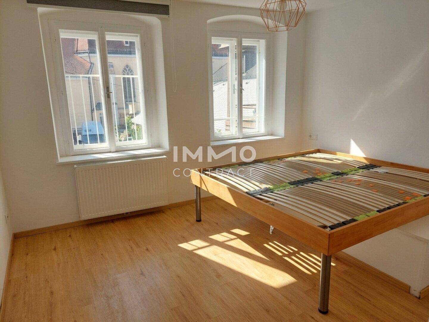 Helles Schlafzimmer mit Holzbettrahmen, zwei Fenstern und Laminatboden.