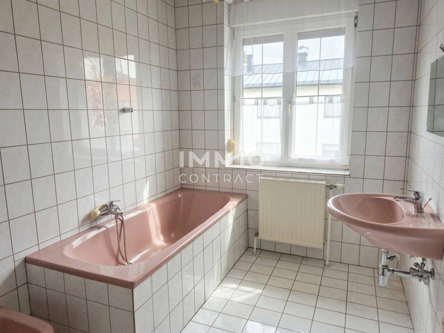 Helles Badezimmer mit weißen Fliesen, rosa Badewanne, Waschbecken und Fenster.