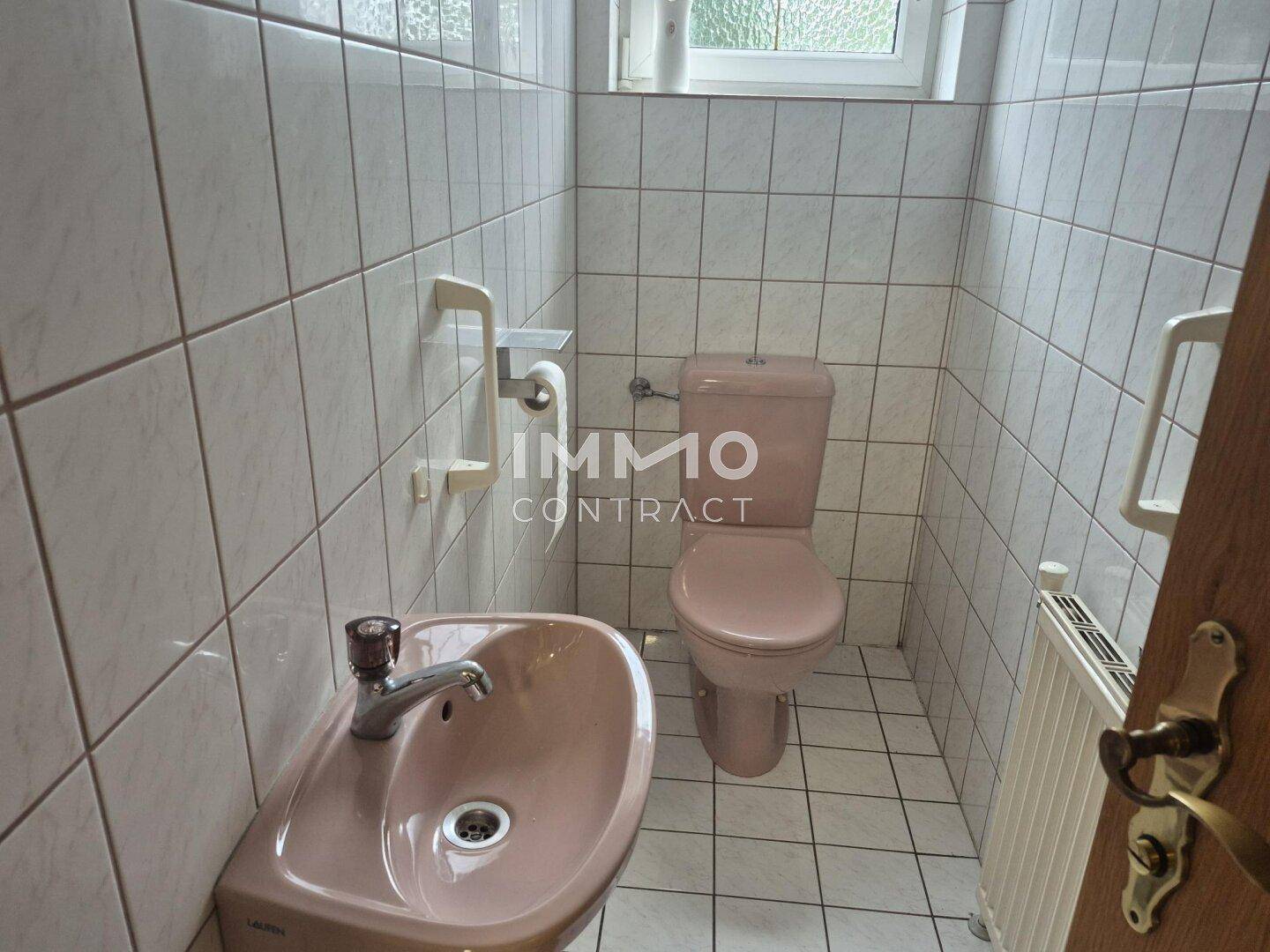 Gäste-WC mit weißen Fliesen, rosa Toilette, Waschbecken und Haltegriffen für Barrierefreiheit.