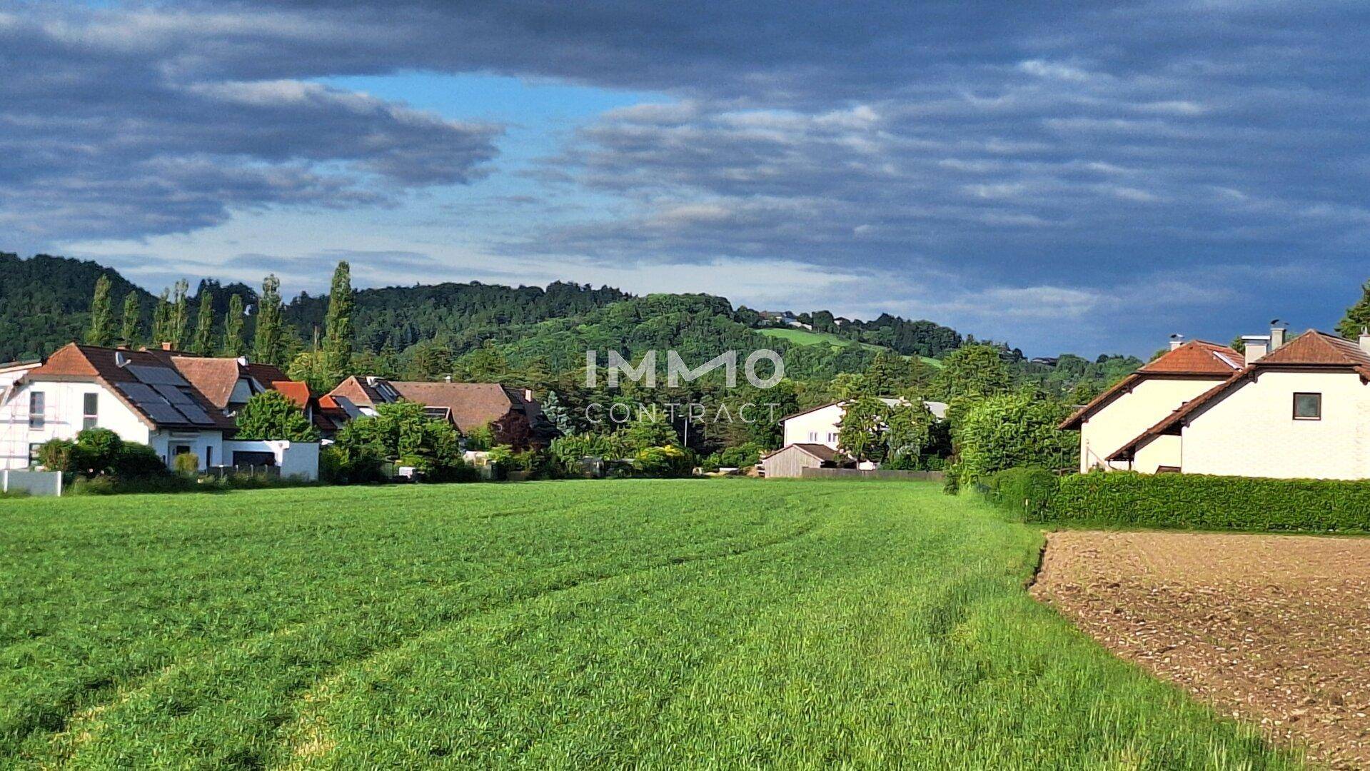 Weites grünes Feld mit ländlicher Bebauung und bewaldeten Hügeln unter bewölktem Himmel.