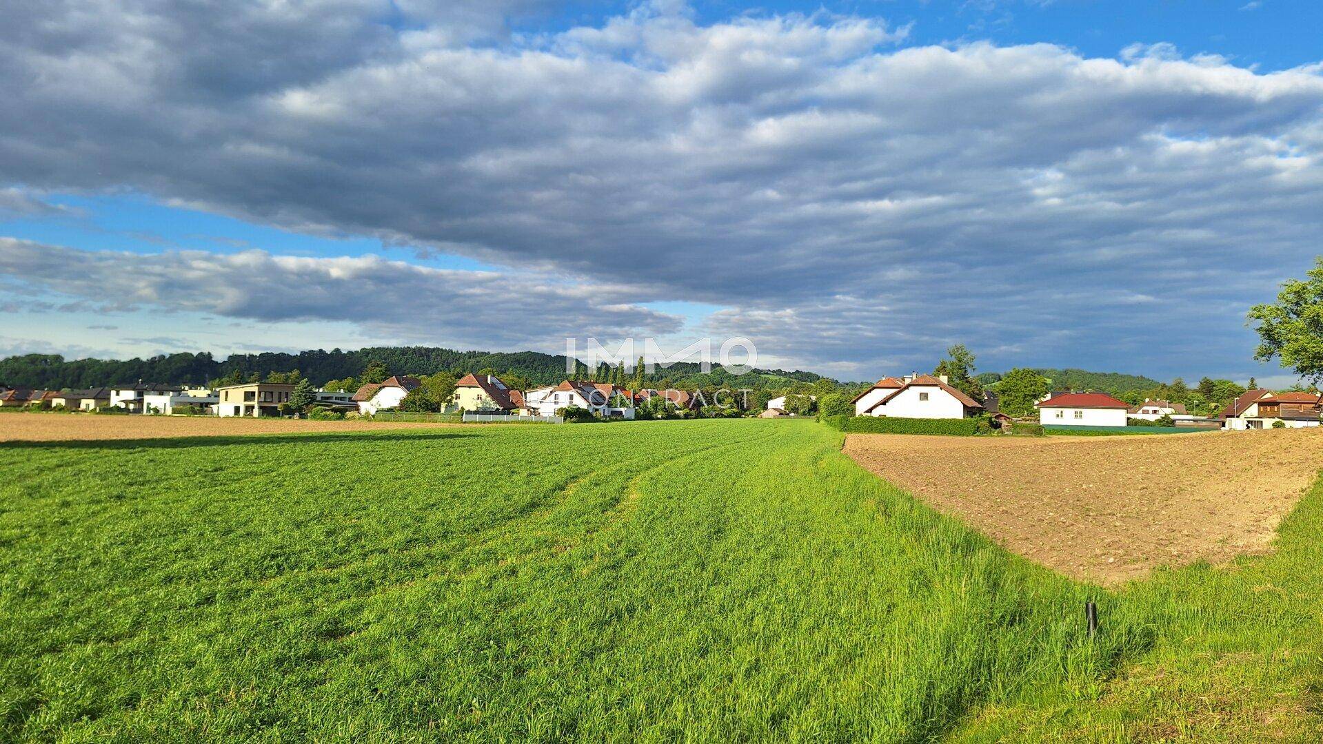 Weitläufiges Feld mit ländlicher Bebauung und bewaldeten Hügeln unter einem bewölkten Himmel.