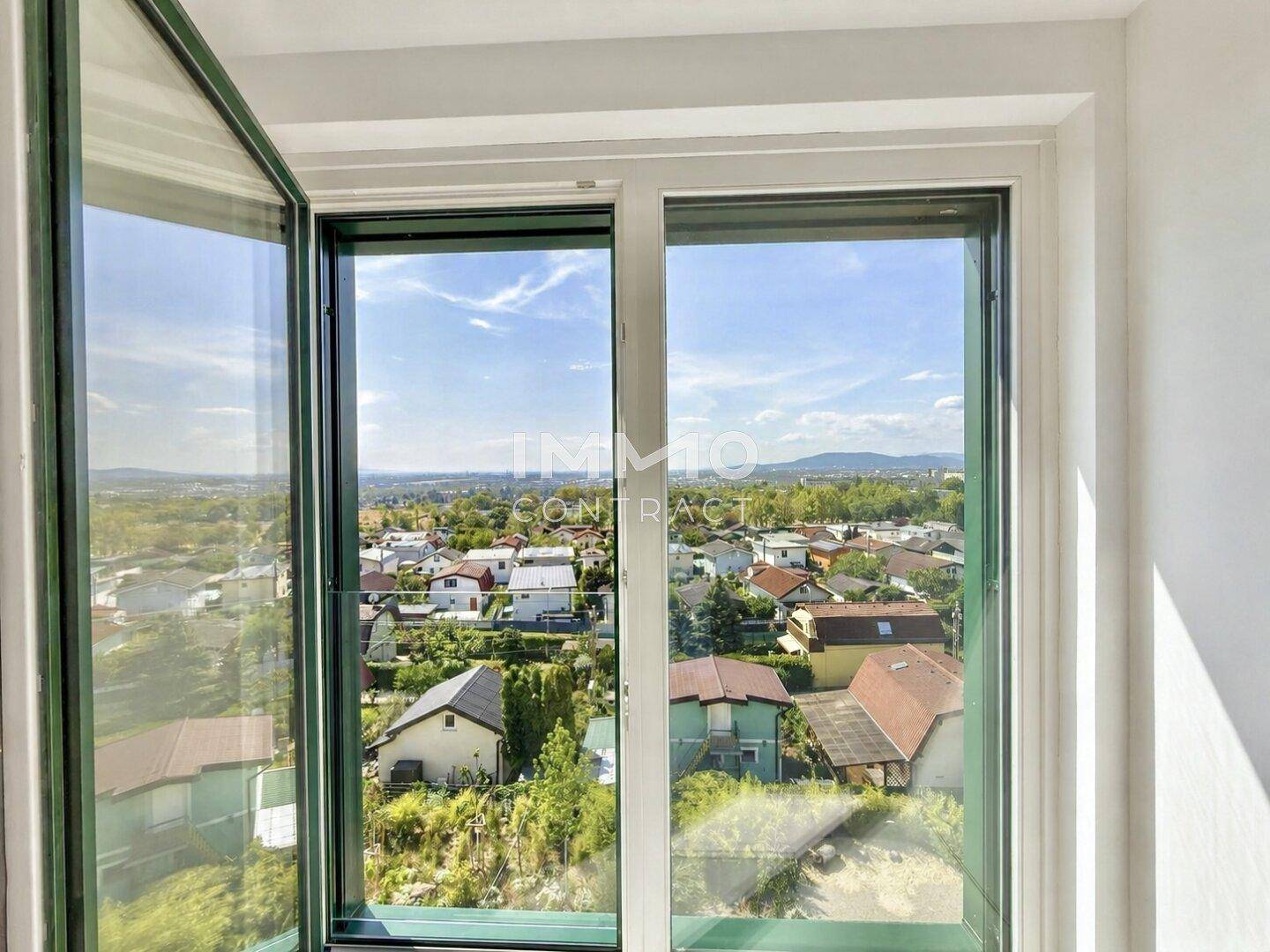 Blick aus einem geöffneten Fenster auf eine Wohnsiedlung mit grünen Bäumen und Bergen in der Ferne.