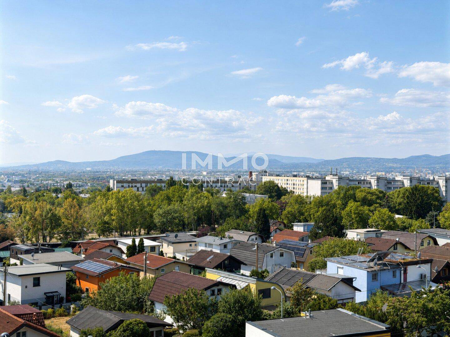Weitläufiger Blick über eine grüne Wohnsiedlung mit vielen Bäumen und Bergen im Hintergrund unter blauem Himmel.