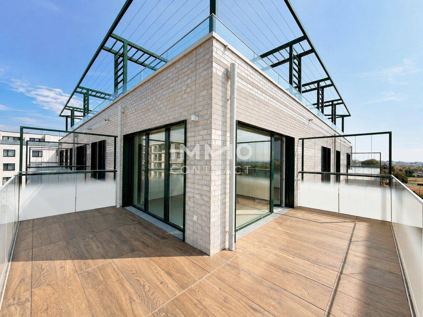 Große Eckterrasse mit Holzboden und Glasgeländer, umgeben von modernen Fenstern und einer hellen Ziegelfassade.