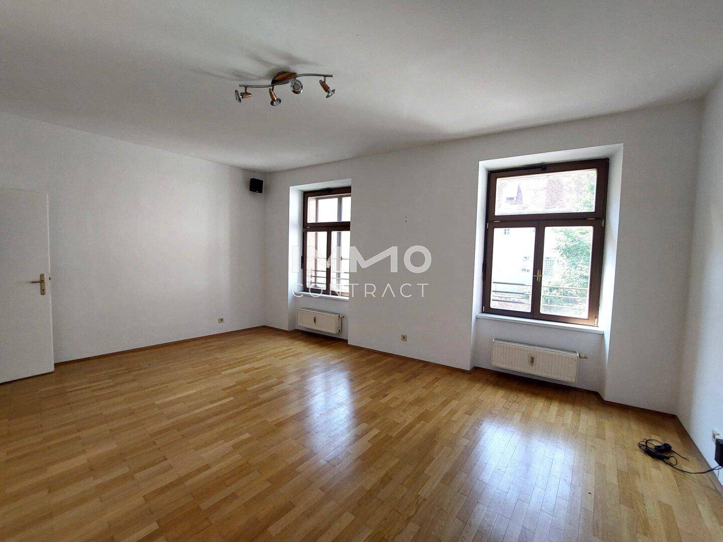Helles, leeres Zimmer mit Parkettboden, weißen Wänden und zwei Fenstern.