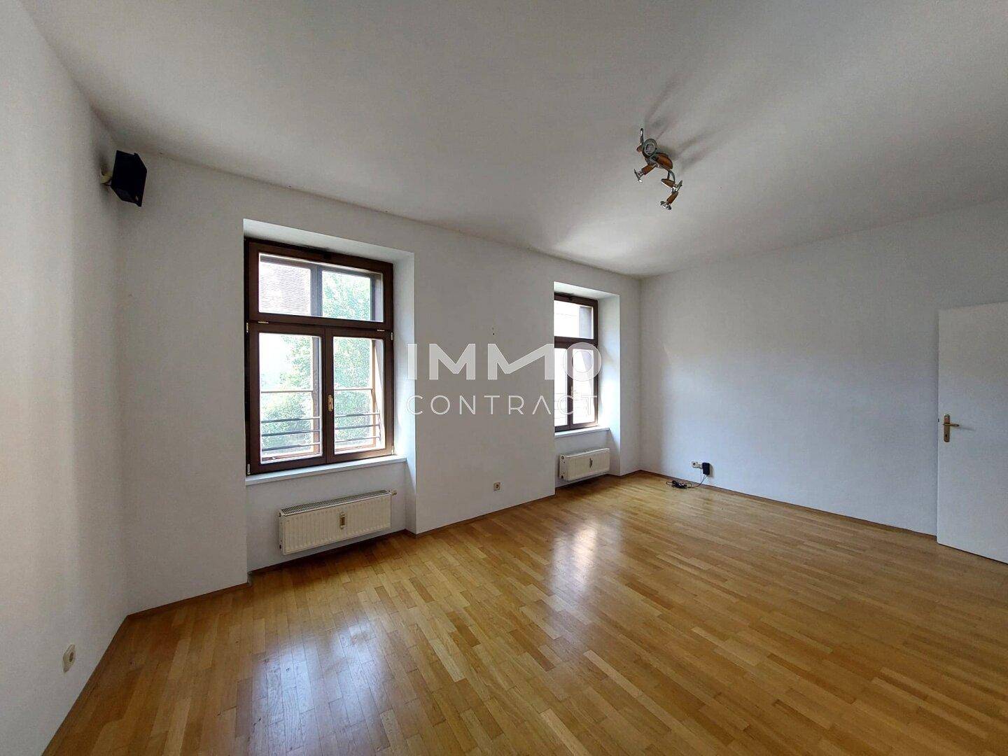Geräumiges Zimmer mit hellem Parkettboden, weißen Wänden und zwei großen Fenstern.