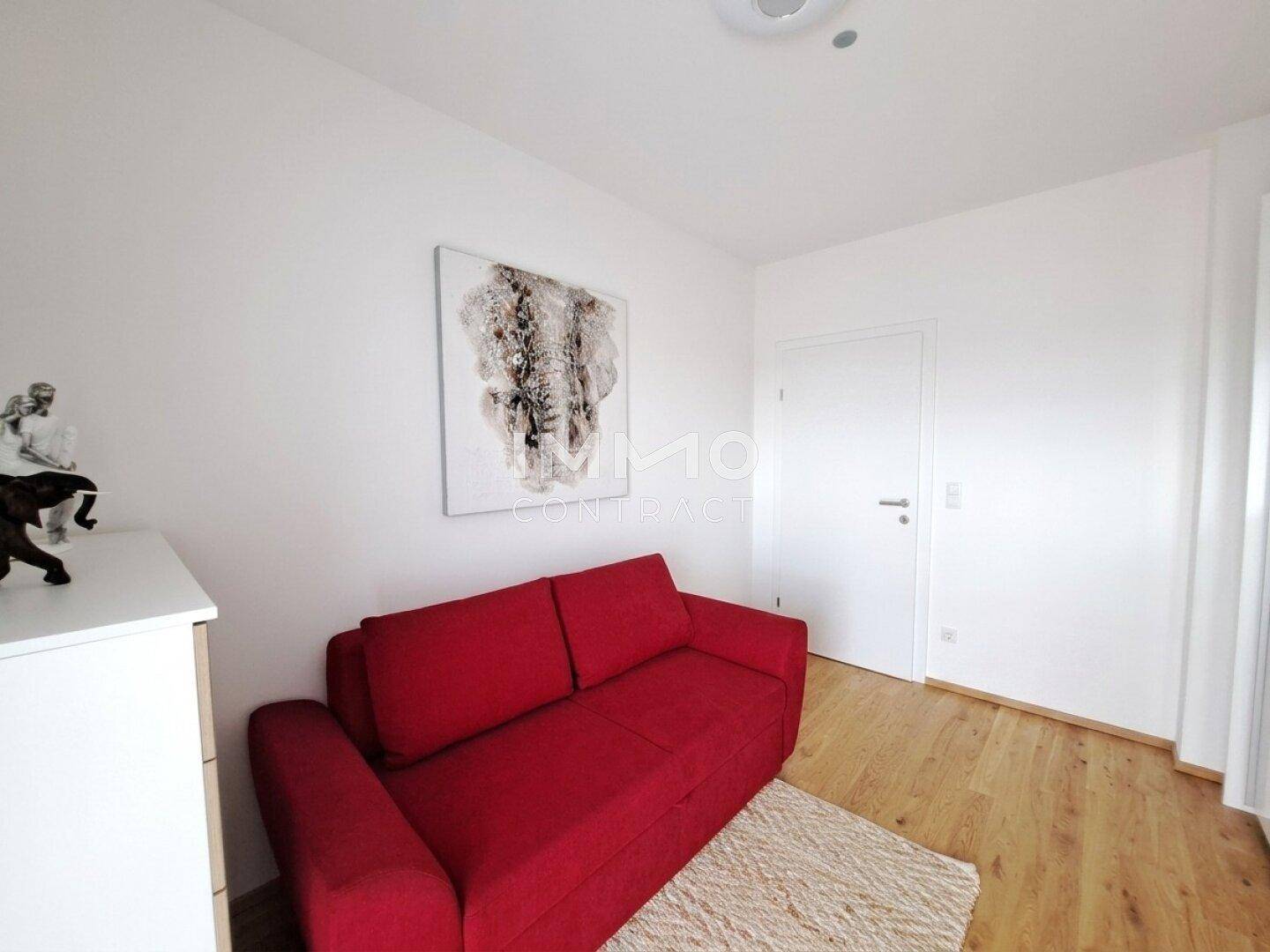 Gemütliches Zimmer mit rotem Sofa, Holzboden und weiß gestrichenen Wänden.