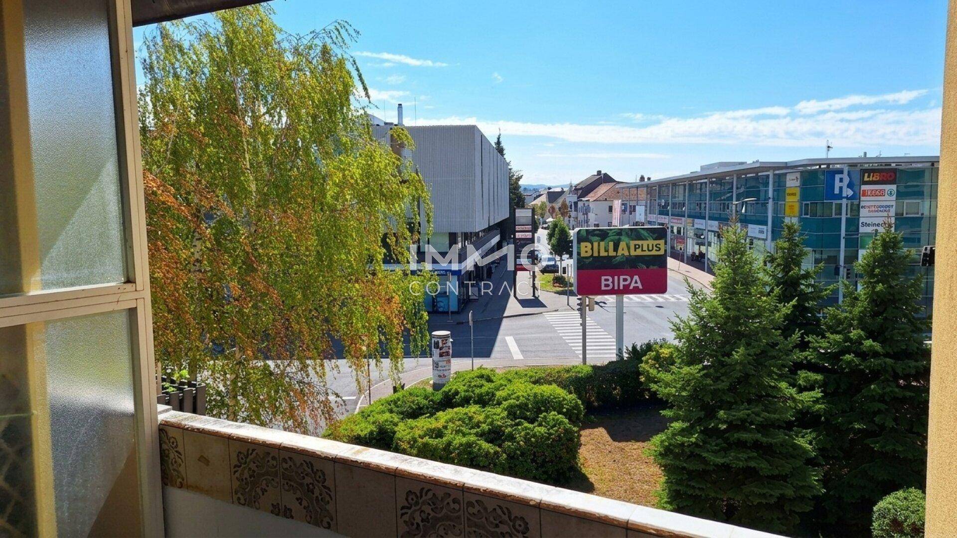 Blick vom Balkon auf eine belebte Straße mit Geschäften und grünen Bäumen.