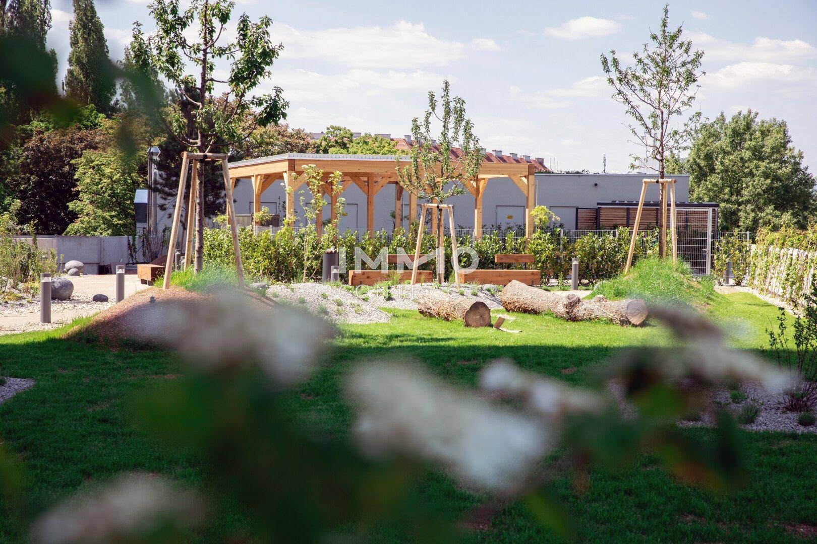 Weitläufige Gemeinschaftsgartenanlage mit Holzpergola, Rasenflächen und jungen Bäumen.
