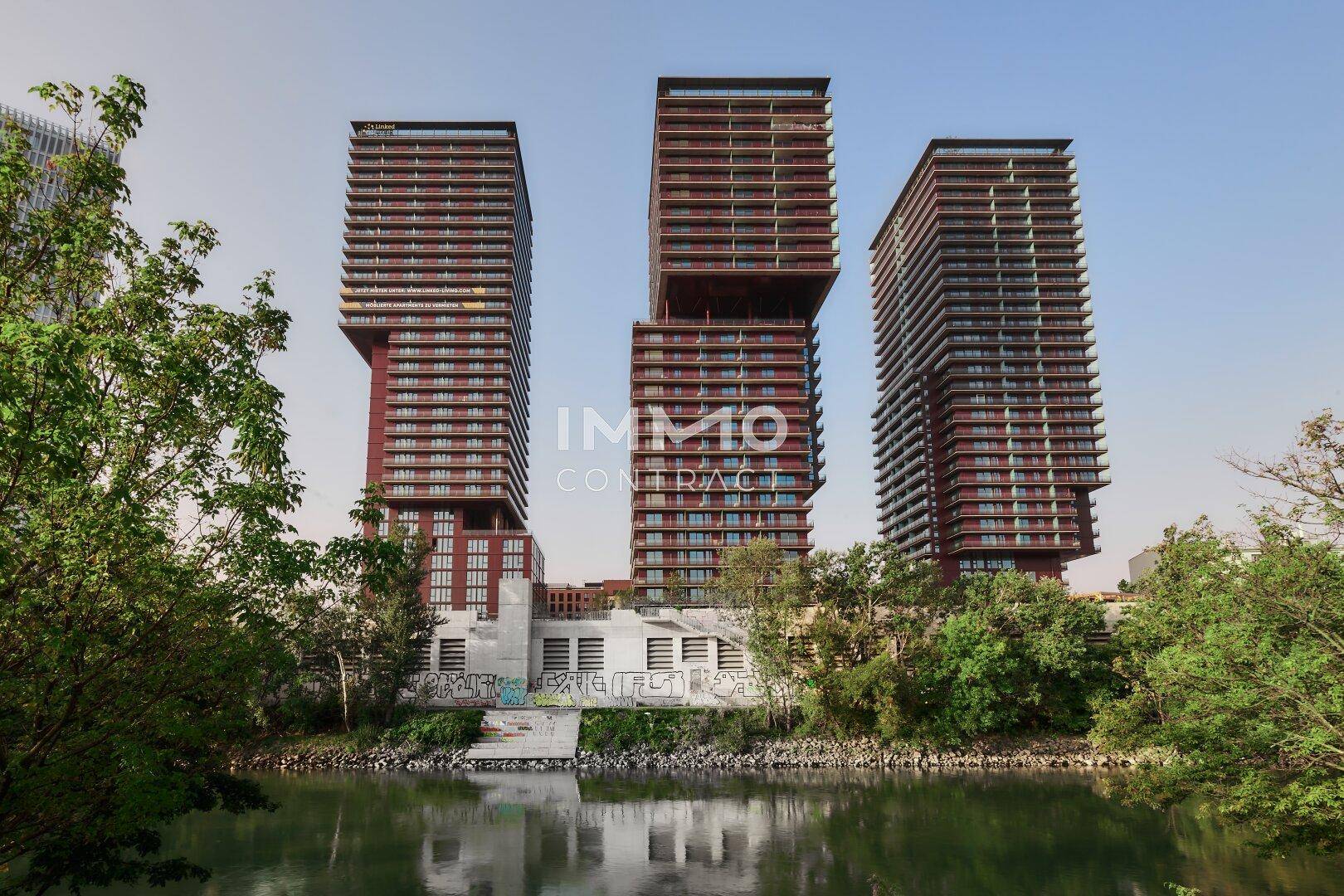 Moderne Hochhäuser mit markanter Architektur und roten Fassaden, umgeben von Grünflächen und Wasser.