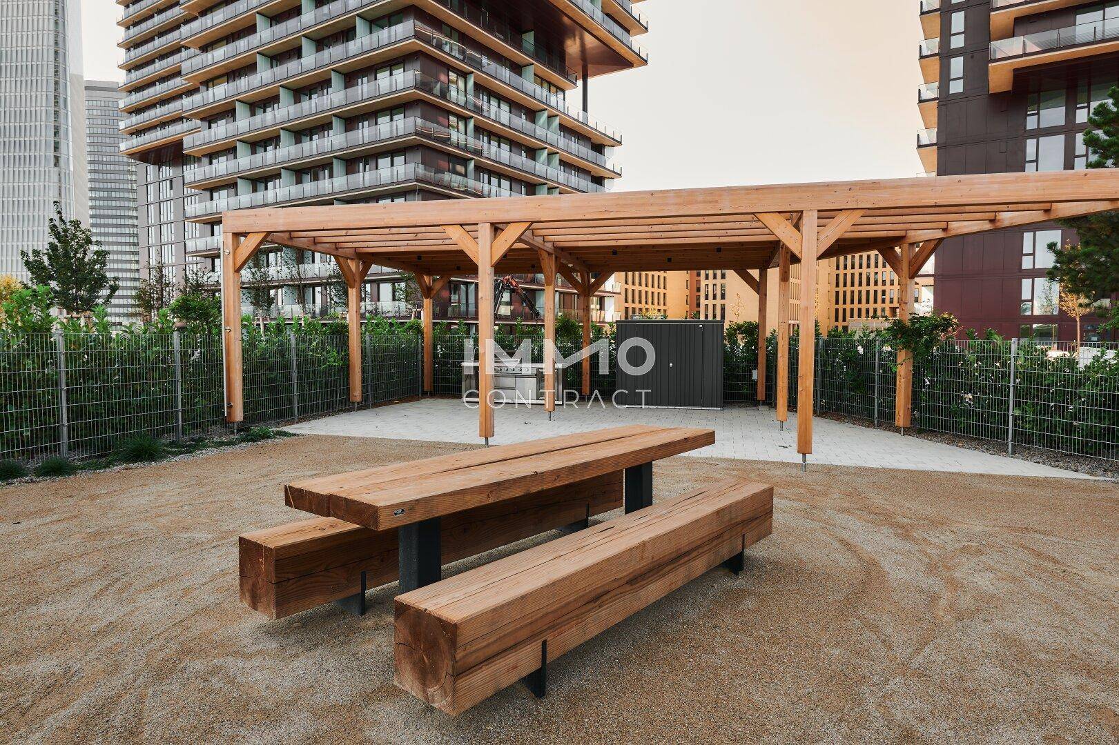 Gemeinschaftsbereich im Freien mit Holztisch und Bänken unter einer Pergola, umgeben von Gebäuden.