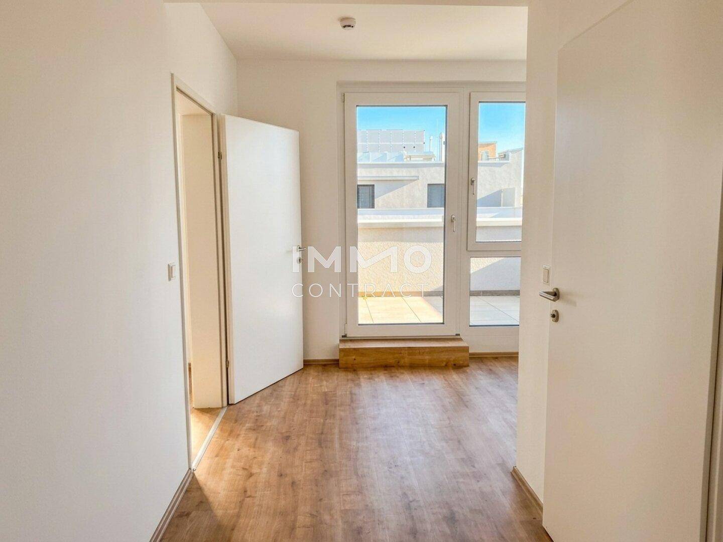 Leeres Zimmer mit Holzboden und großem Fenster, das viel Tageslicht hereinlässt.
