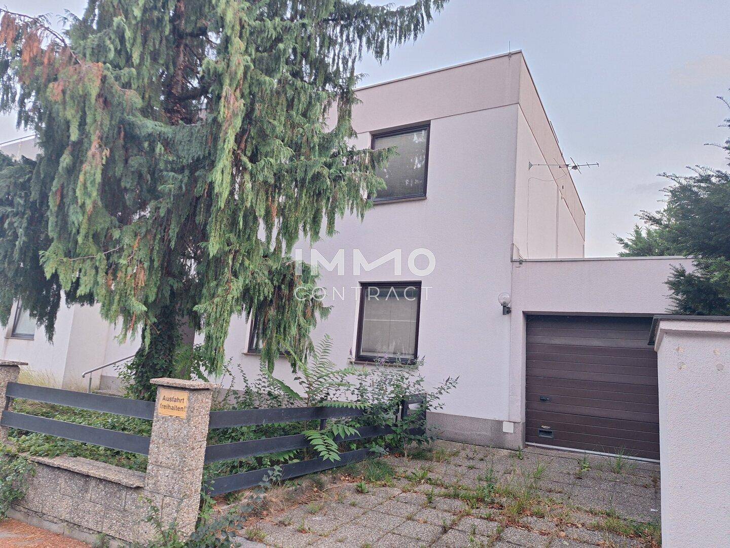 Seitenansicht des Hauses mit Garagentor, Zaun und einem großen Baum im Vordergrund.