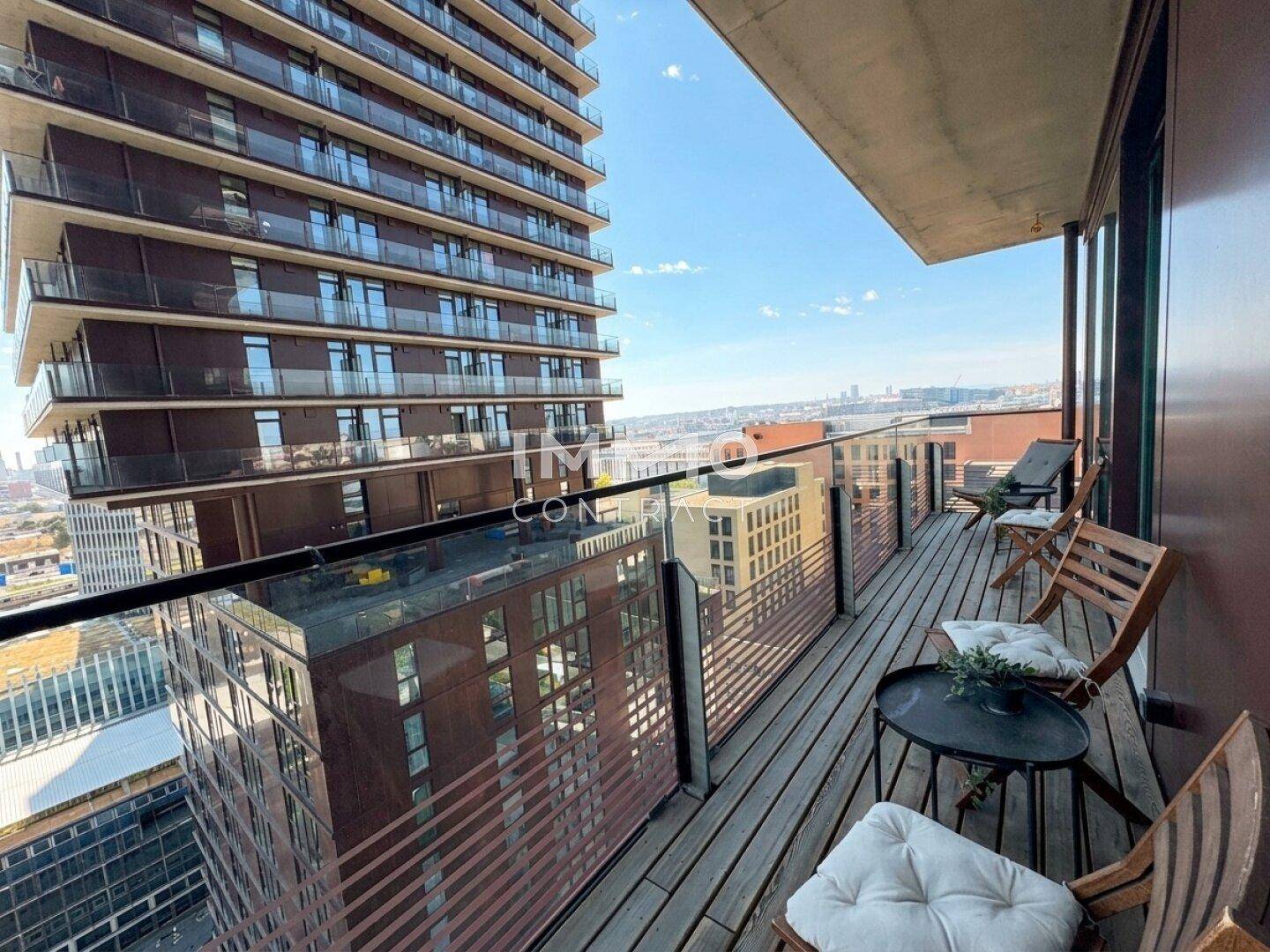Geräumiger Balkon mit Holzdeck und Glasgeländer, umgeben von moderner Hochhausarchitektur.