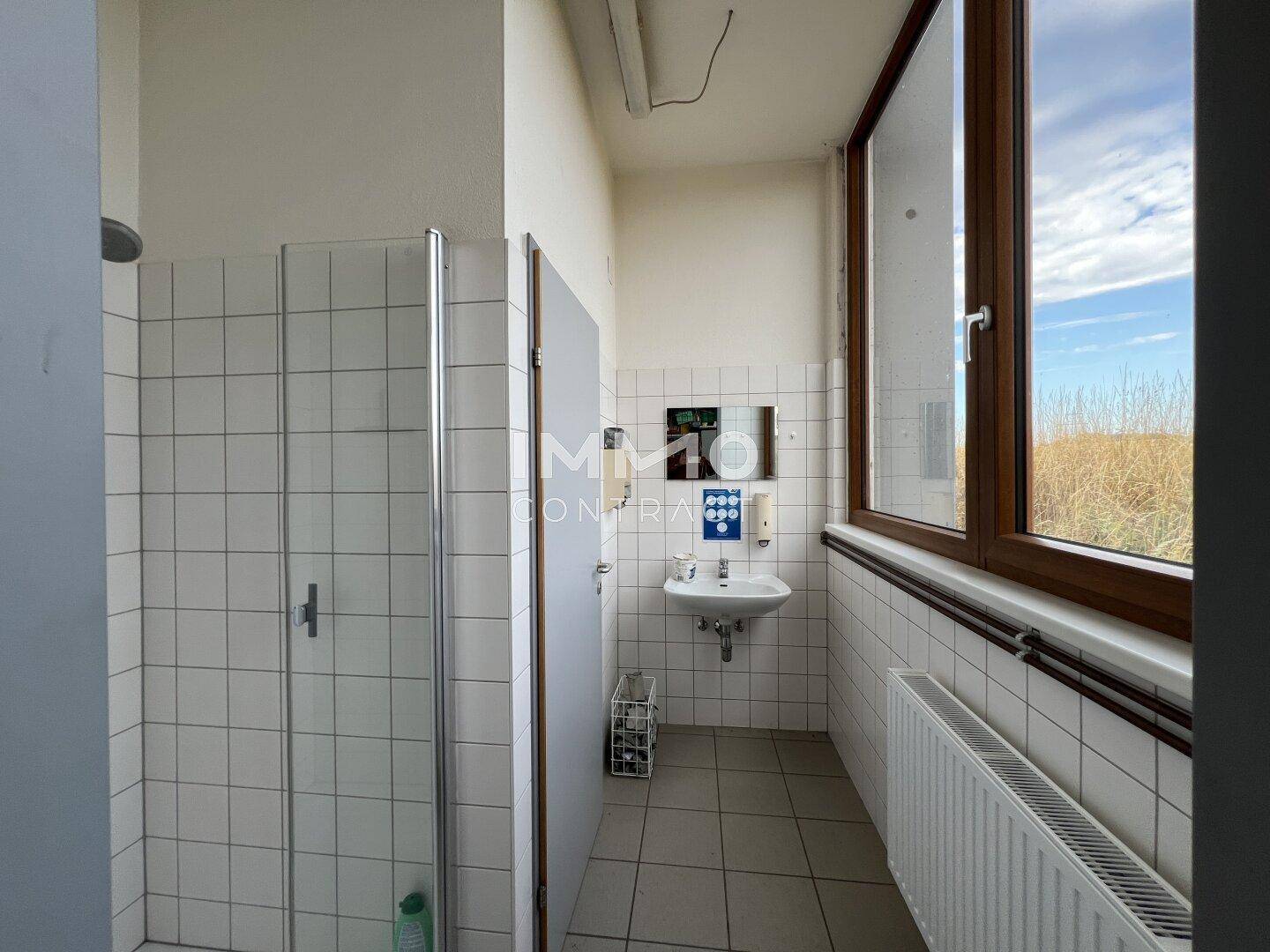 Einfaches Badezimmer mit Duschkabine, Waschbecken und Fenster, gefliest in Weiß und Hellgrau.