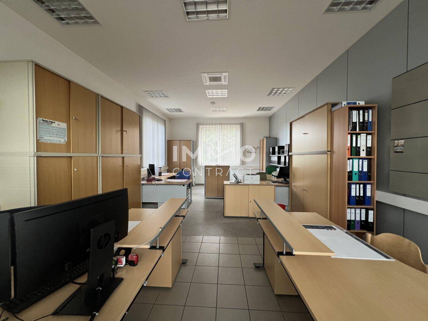 Geräumiges Büro mit Holzschränken, mehreren Schreibtischen und Fliesenboden, gut beleuchtet durch Fenster.
