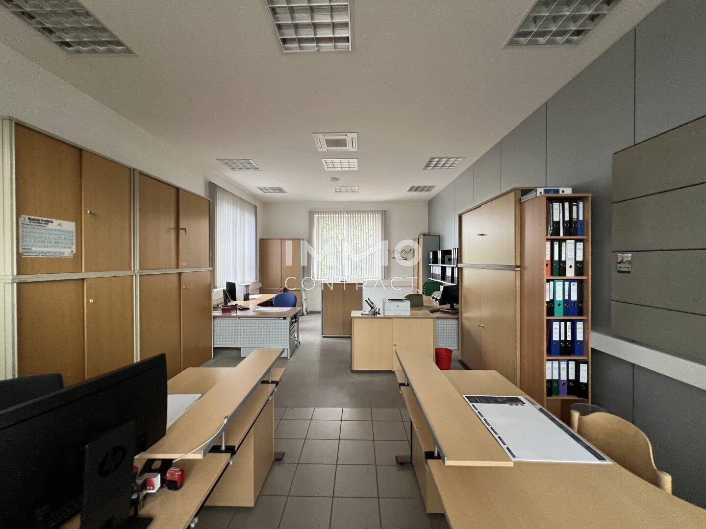 Geräumiges Büro mit Holzschränken, mehreren Schreibtischen und Fliesenboden, gut beleuchtet durch Fenster.