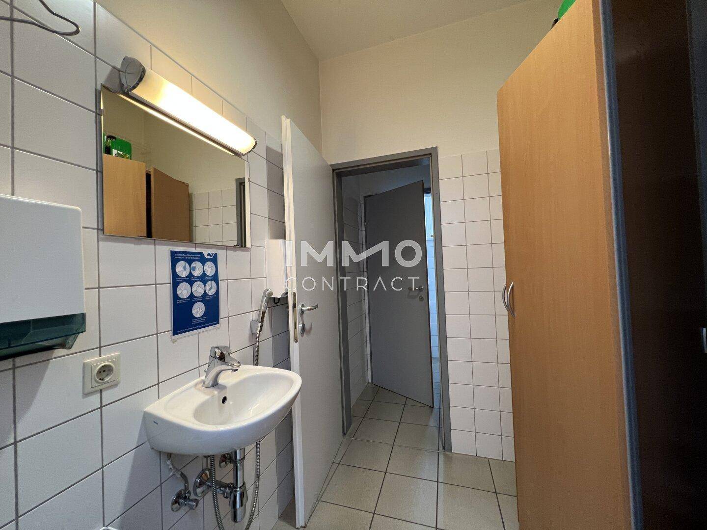 Badezimmer mit Waschbecken, Spiegel und weiß gefliesten Wänden, Zugang zu einer Toilette.