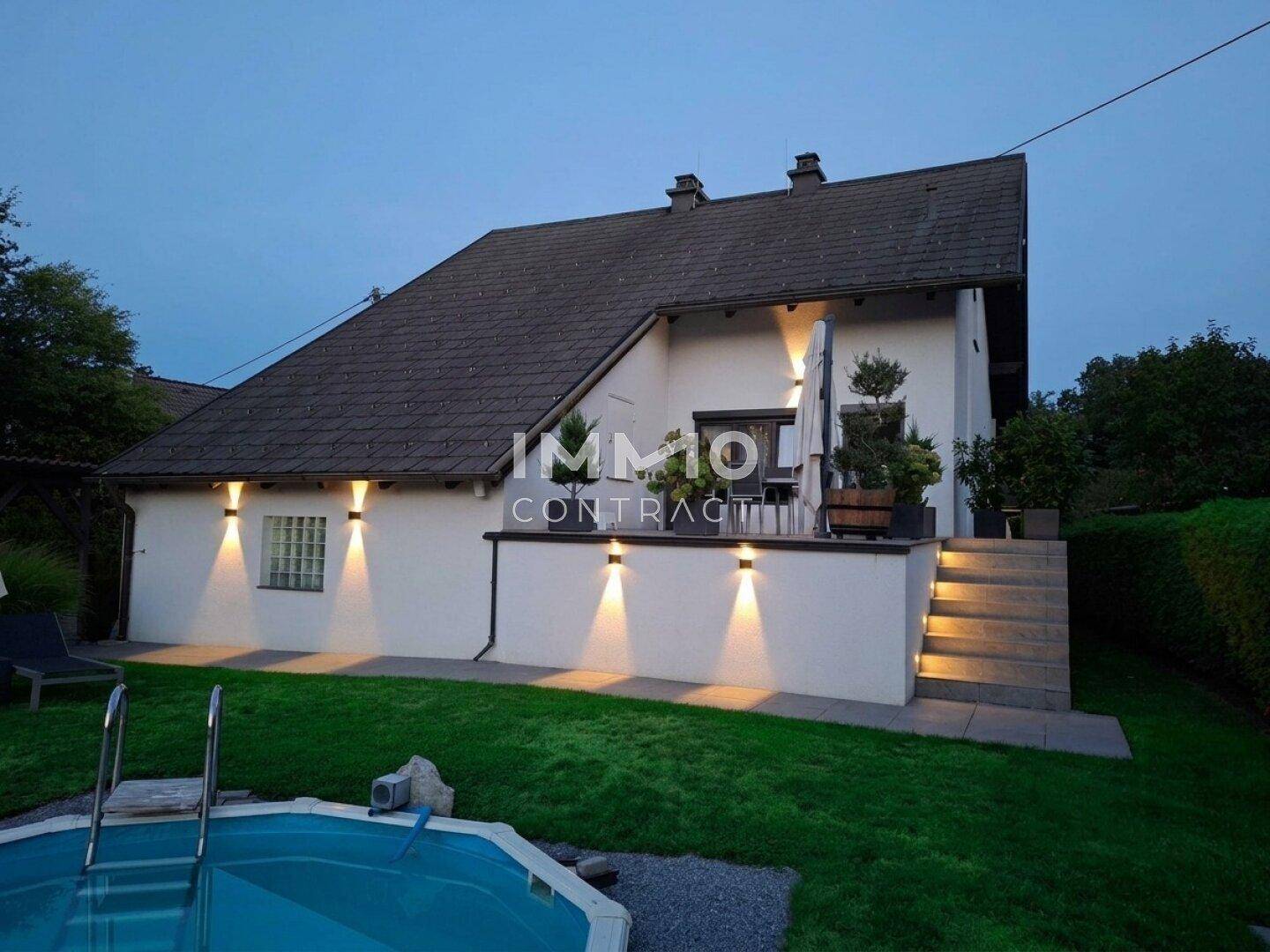 Hausfassade bei Dämmerung mit beleuchteter Terrasse und Gartenbereich.