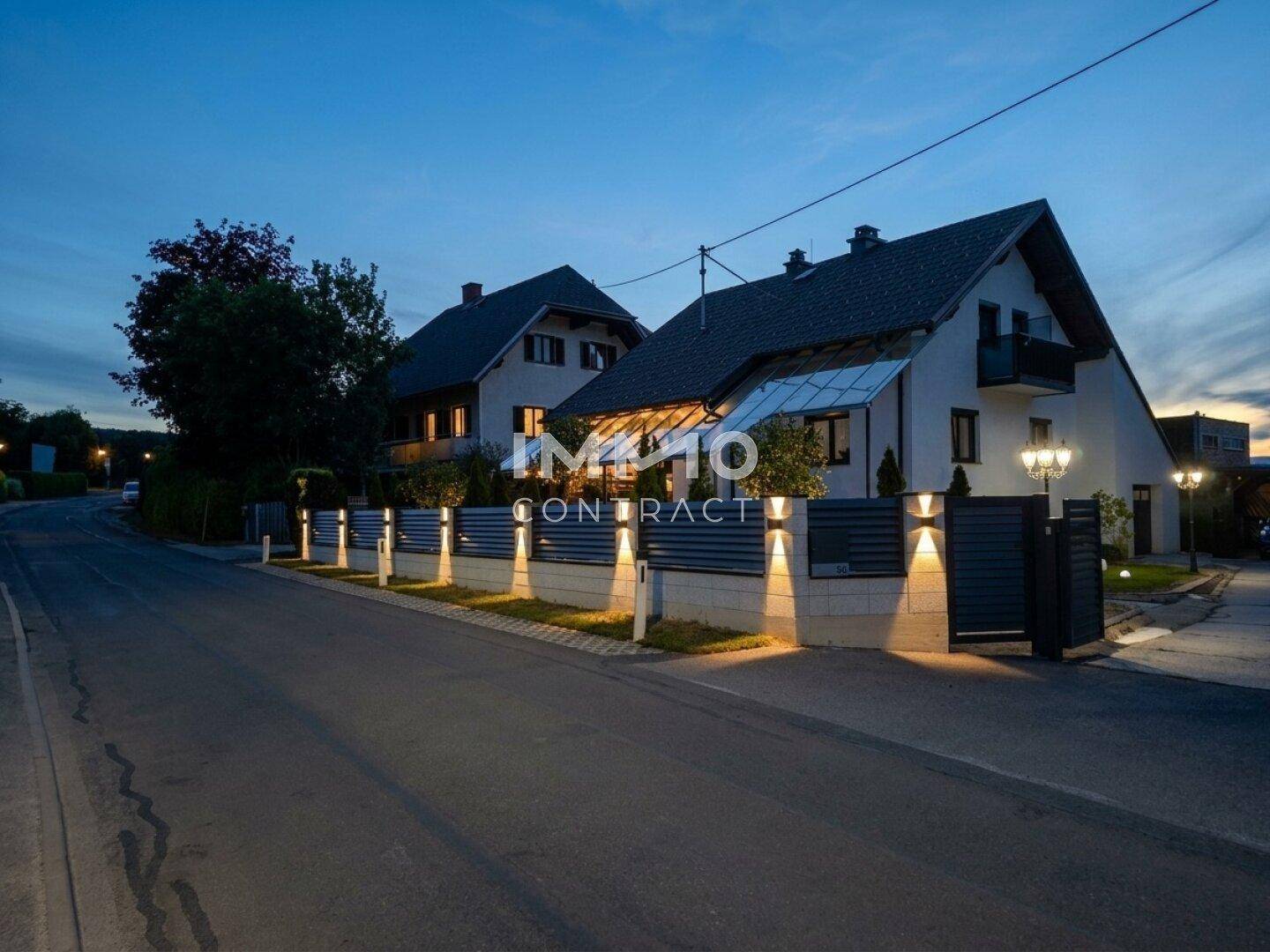 Außenansicht des Hauses und der modernen Zaunanlage bei Dämmerung mit Beleuchtung.