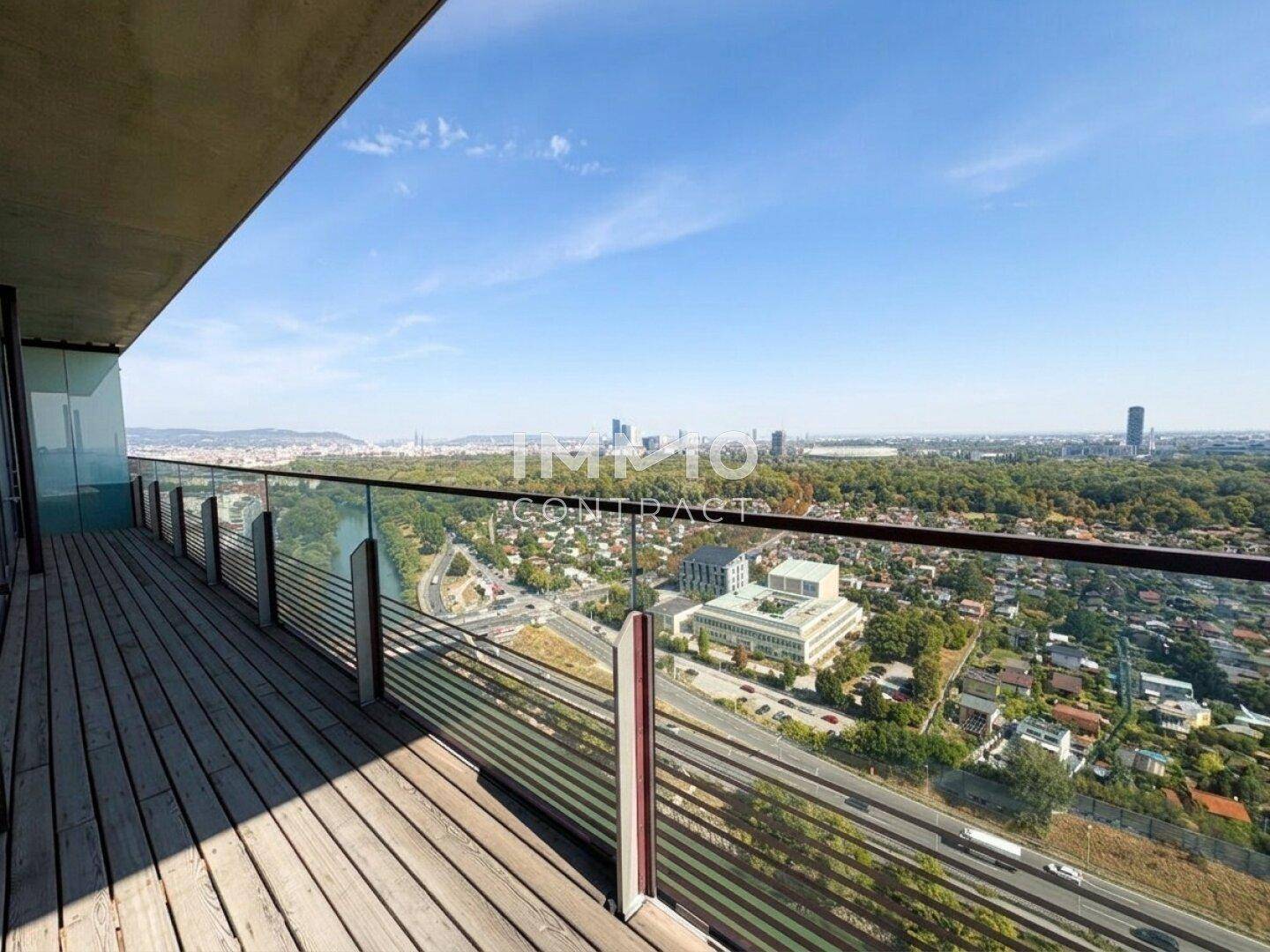 Geräumiger Balkon mit Holzdeck und Glasgeländer bietet weite Aussicht auf Stadt und Grünflächen.