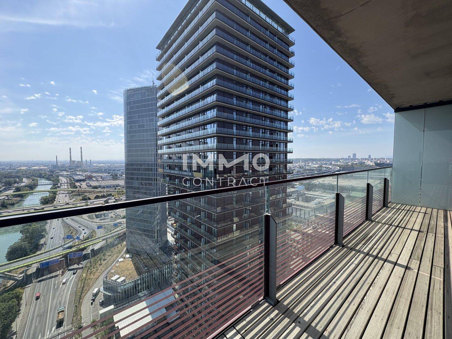 Balkon mit Holzdeck und Glasgeländer bietet Blick auf ein modernes Hochhaus und die Umgebung.