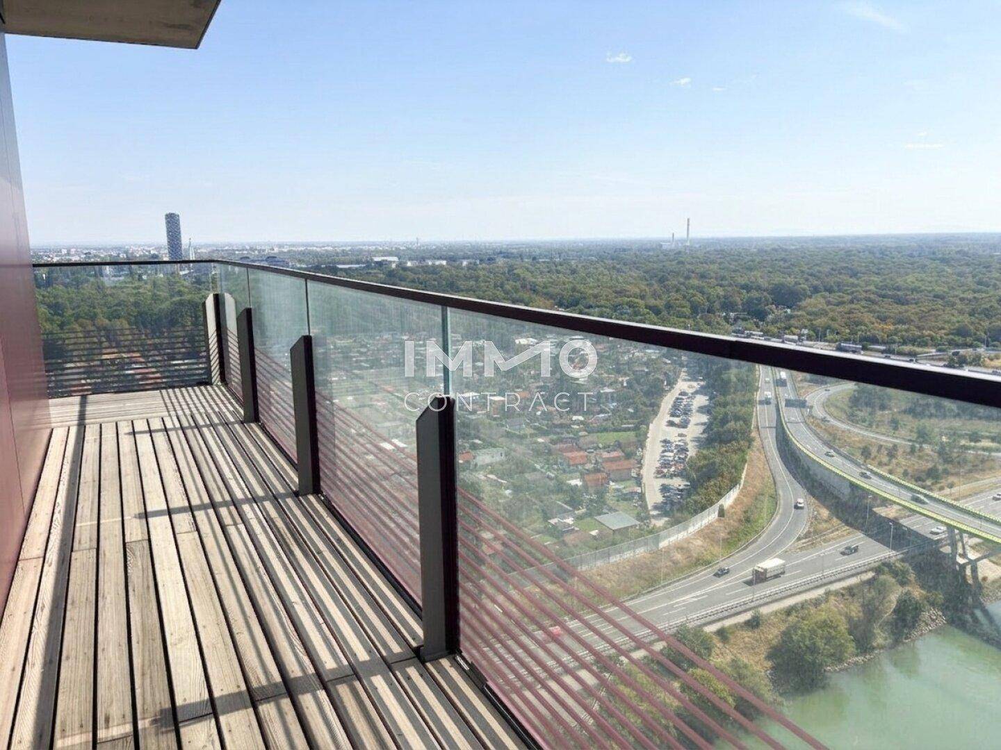 Geräumiger Balkon mit Holzbelag und Glasgeländer bietet weite Aussicht auf Autobahn und Grünflächen.