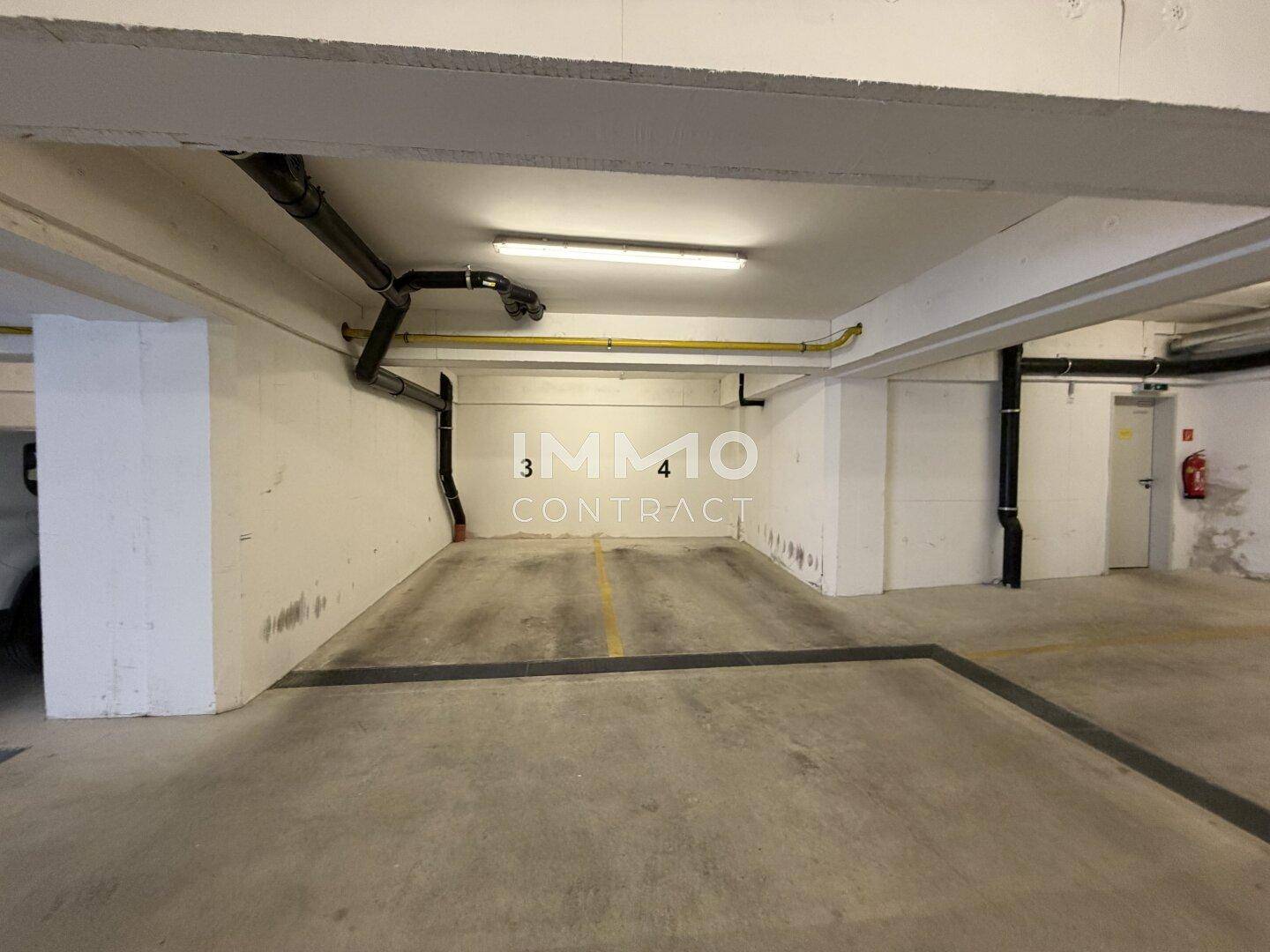 Leerer Parkplatz in einer Tiefgarage mit sichtbaren Rohren und einem Feuerlöscher an der Wand.