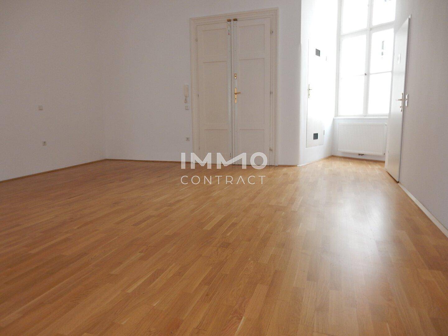Leerer Raum mit hellem Parkettboden, einer Doppeltür und einem hohen Fenster.