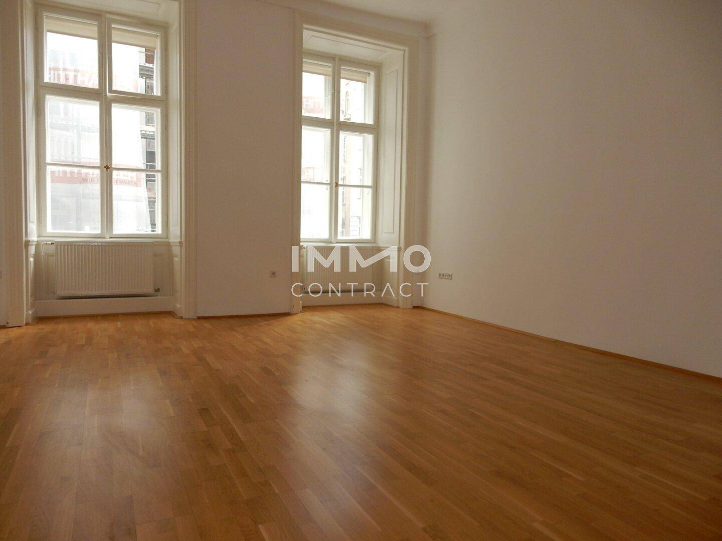 Leerer Raum mit zwei hohen Fenstern, hellem Parkettboden und weißen Wänden.