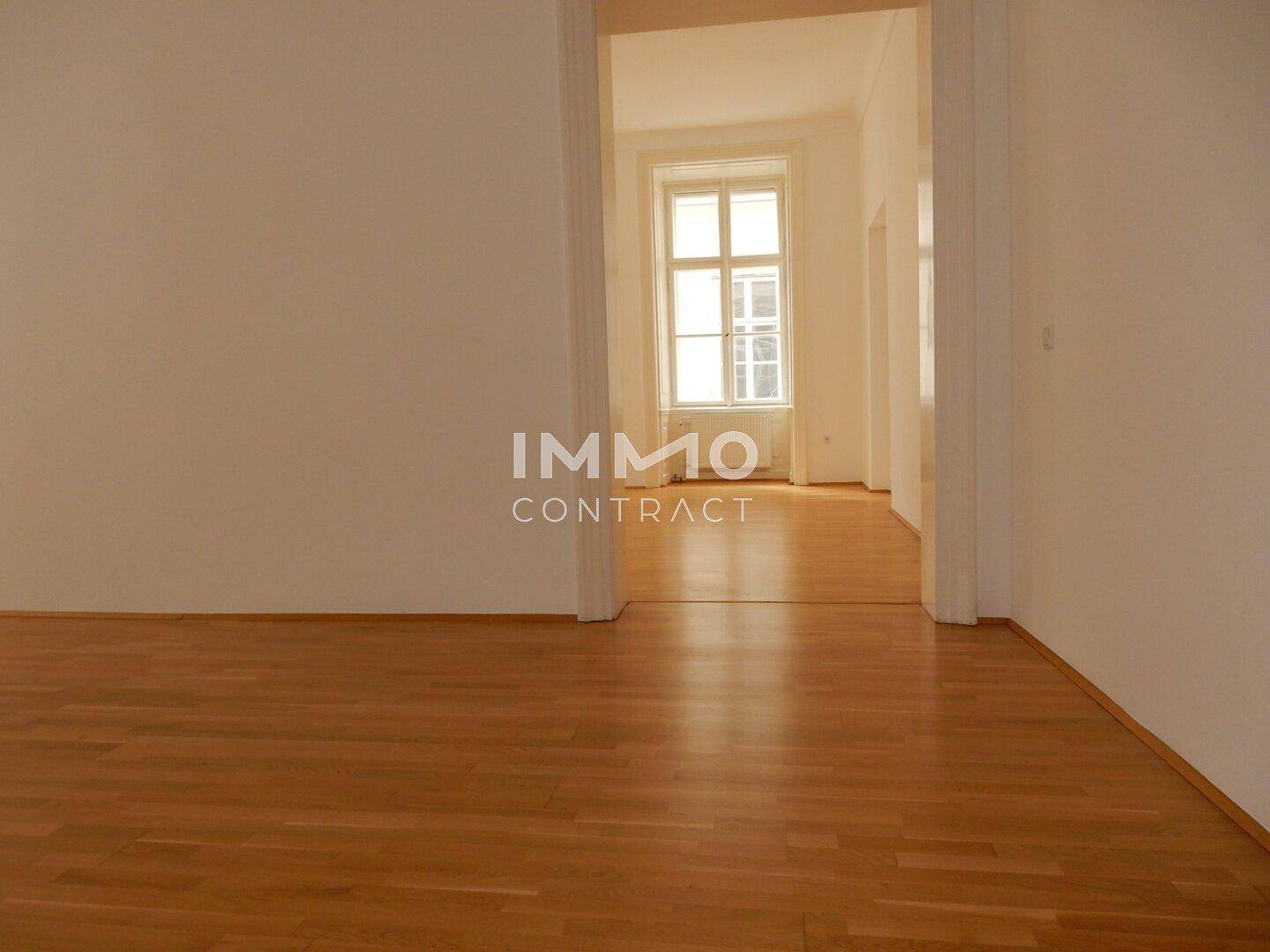 Leerer Raum mit Parkettboden und Durchgang zu einem weiteren hellen Zimmer mit Fenster.