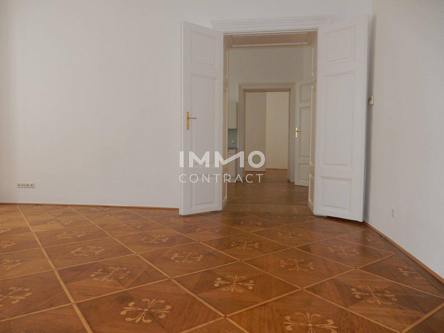 Geräumiger Raum mit gemustertem Parkettboden und offenem Durchgang zu einem weiteren Zimmer.