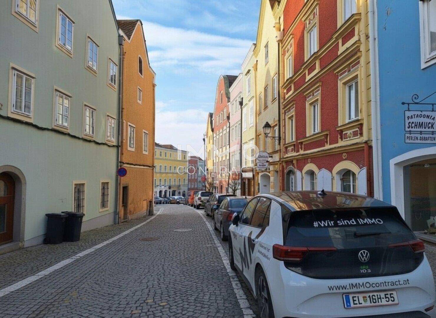 Belebte Kopfsteinpflastergasse mit farbenfrohen historischen Gebäuden und parkenden Autos.