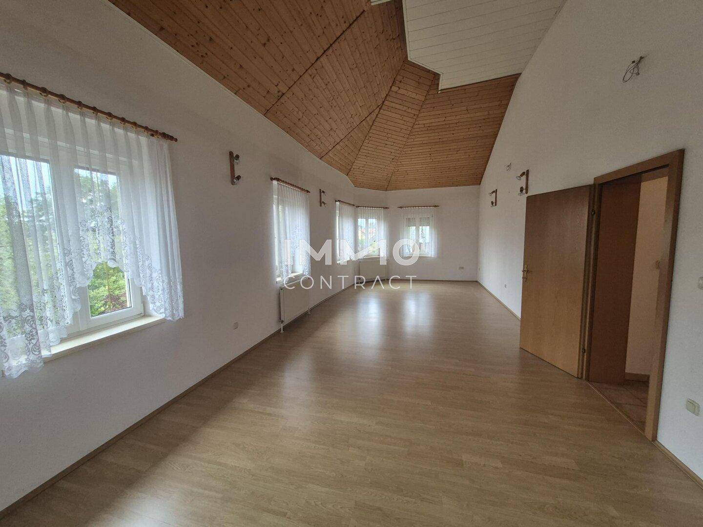 Großer Wohnraum mit Laminatboden, hoher Holzdecke und vielen Fenstern, die den Raum erhellen.