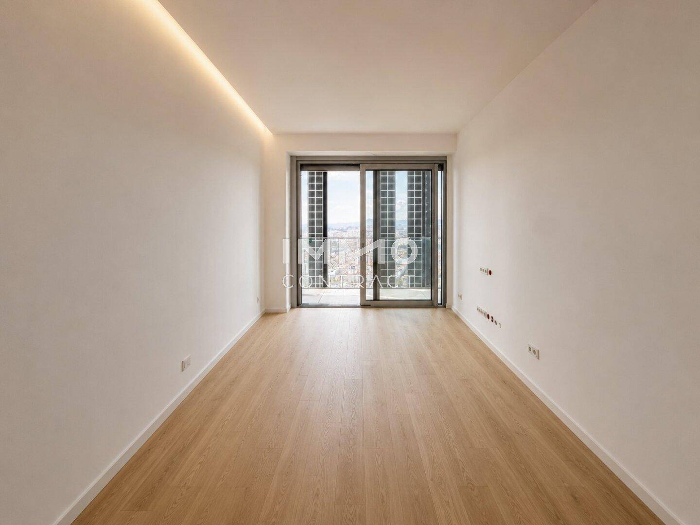 Leeres Zimmer mit Holzfußboden, indirekter Deckenbeleuchtung und bodentiefen Fenstern.