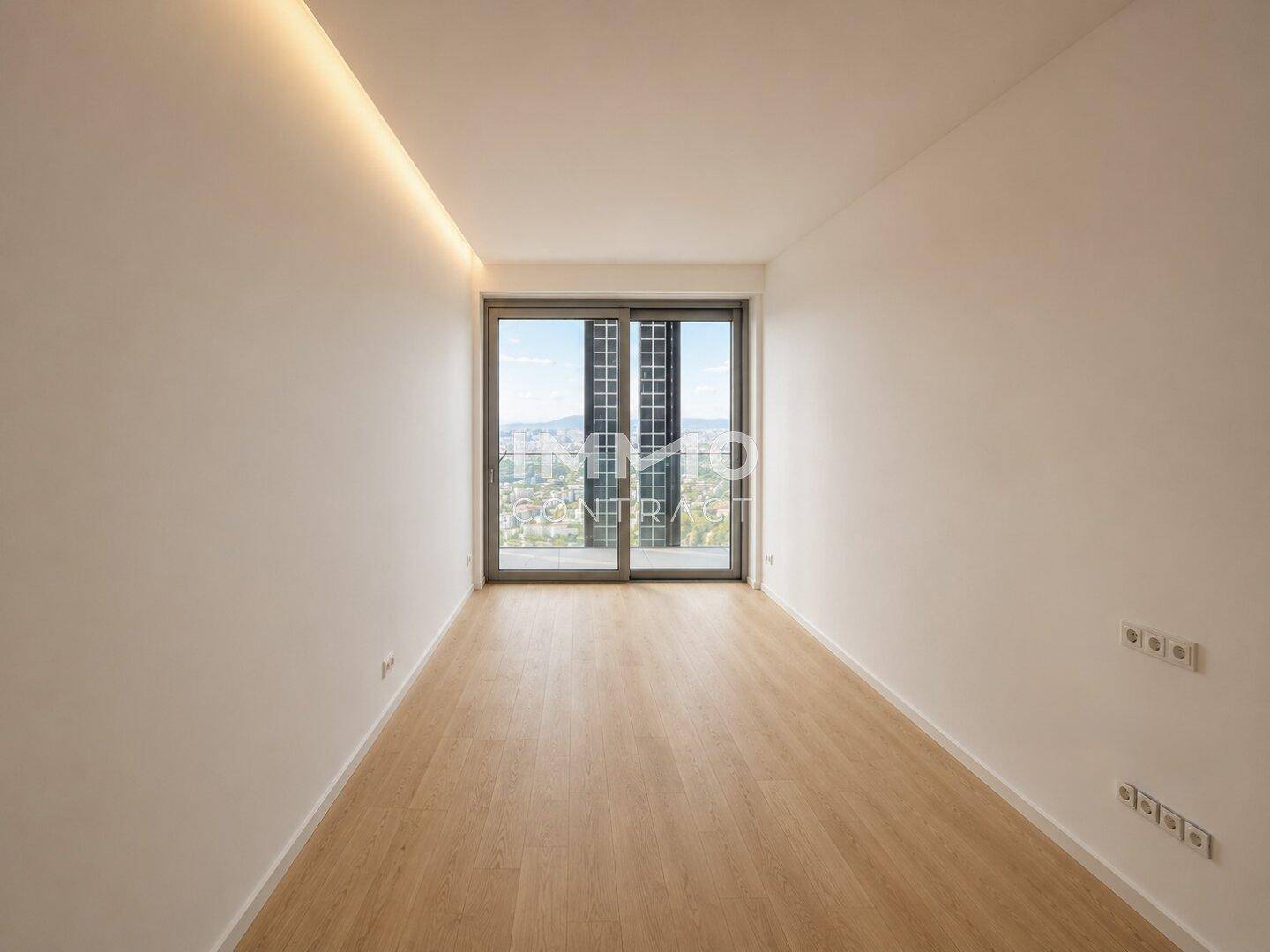 Helles, leeres Zimmer mit Holzfußboden, indirekter Beleuchtung und großen Fenstern mit Aussicht.