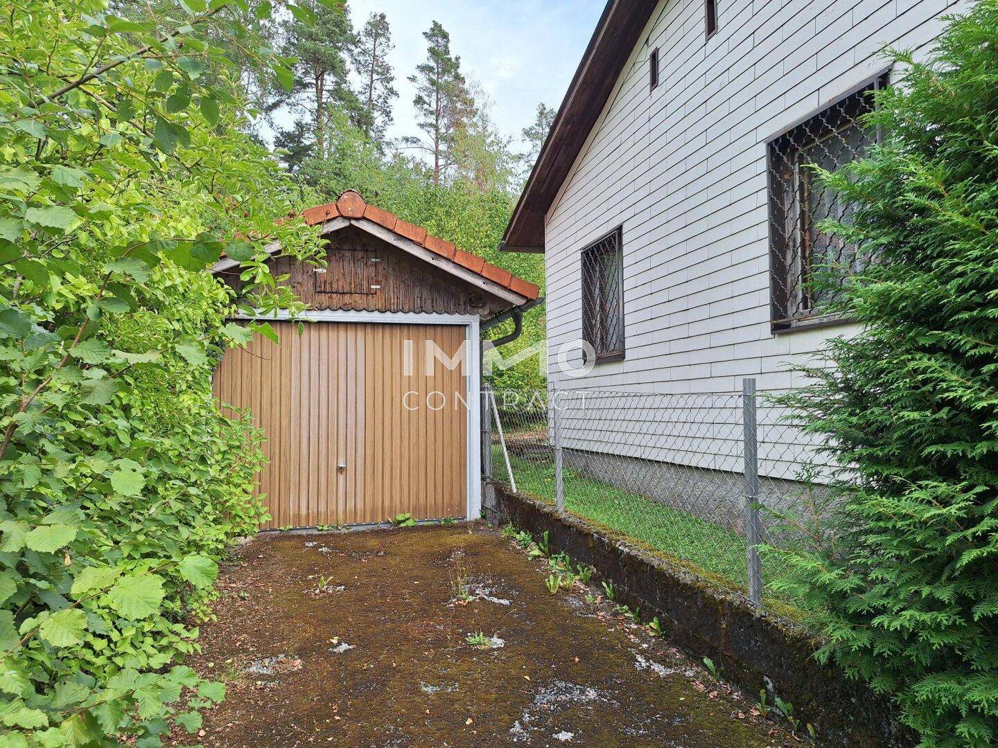 Einzelgarage mit Holztor neben dem Haus, umgeben von üppigem Grün.