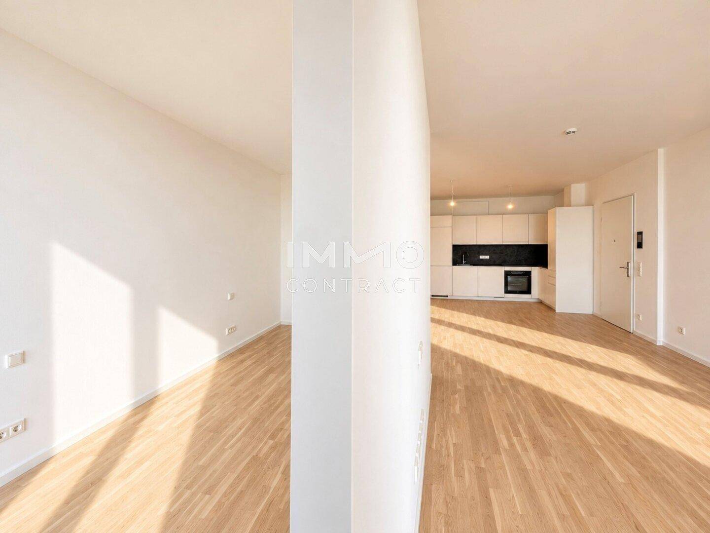 Heller, offener Wohnbereich mit Holzboden und Blick auf eine moderne Einbauküche im Hintergrund.
