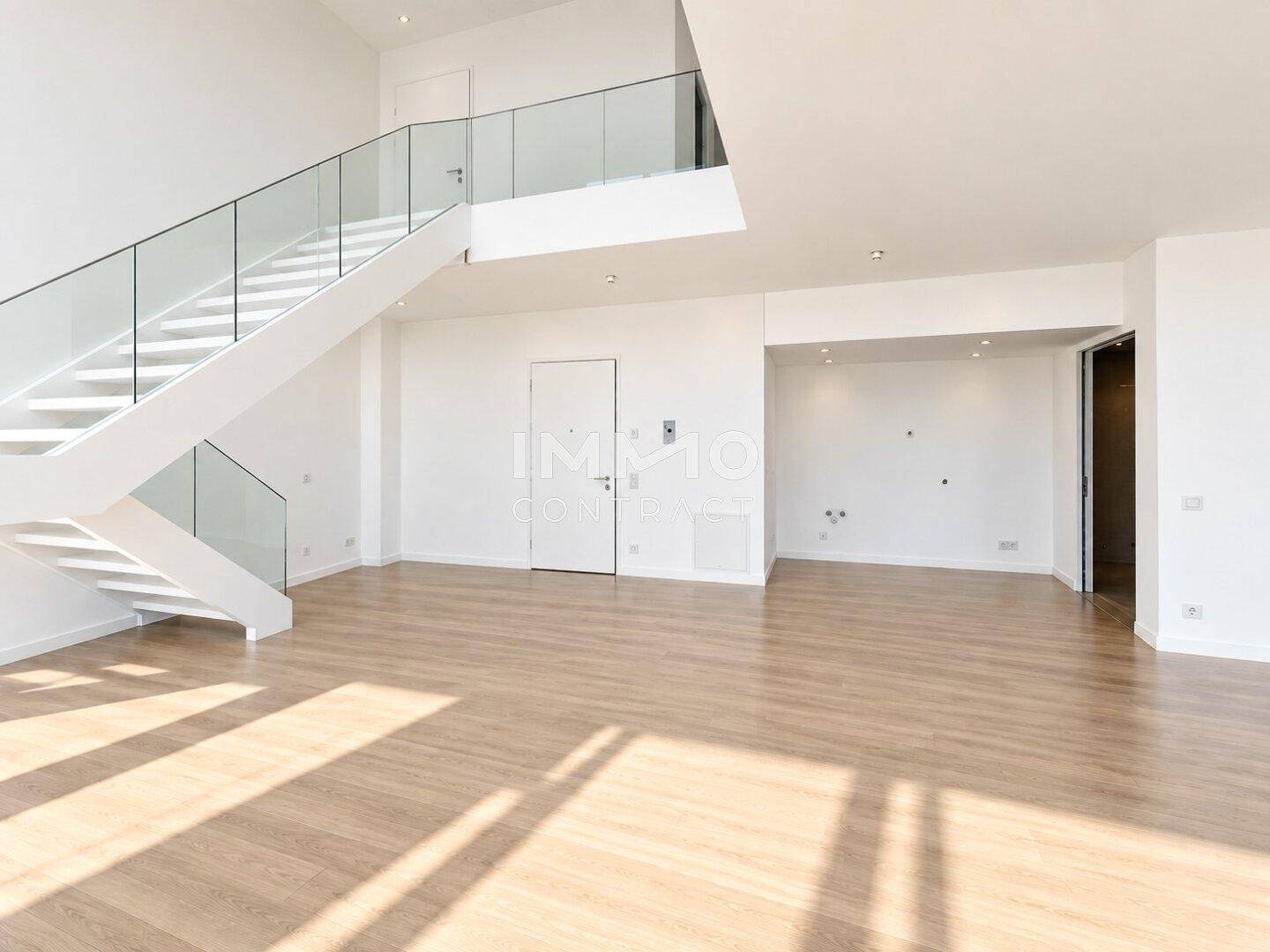 Großer, heller Wohnraum mit Holzfußboden, hohen Decken und einer eleganten Treppe mit Glasgeländer.