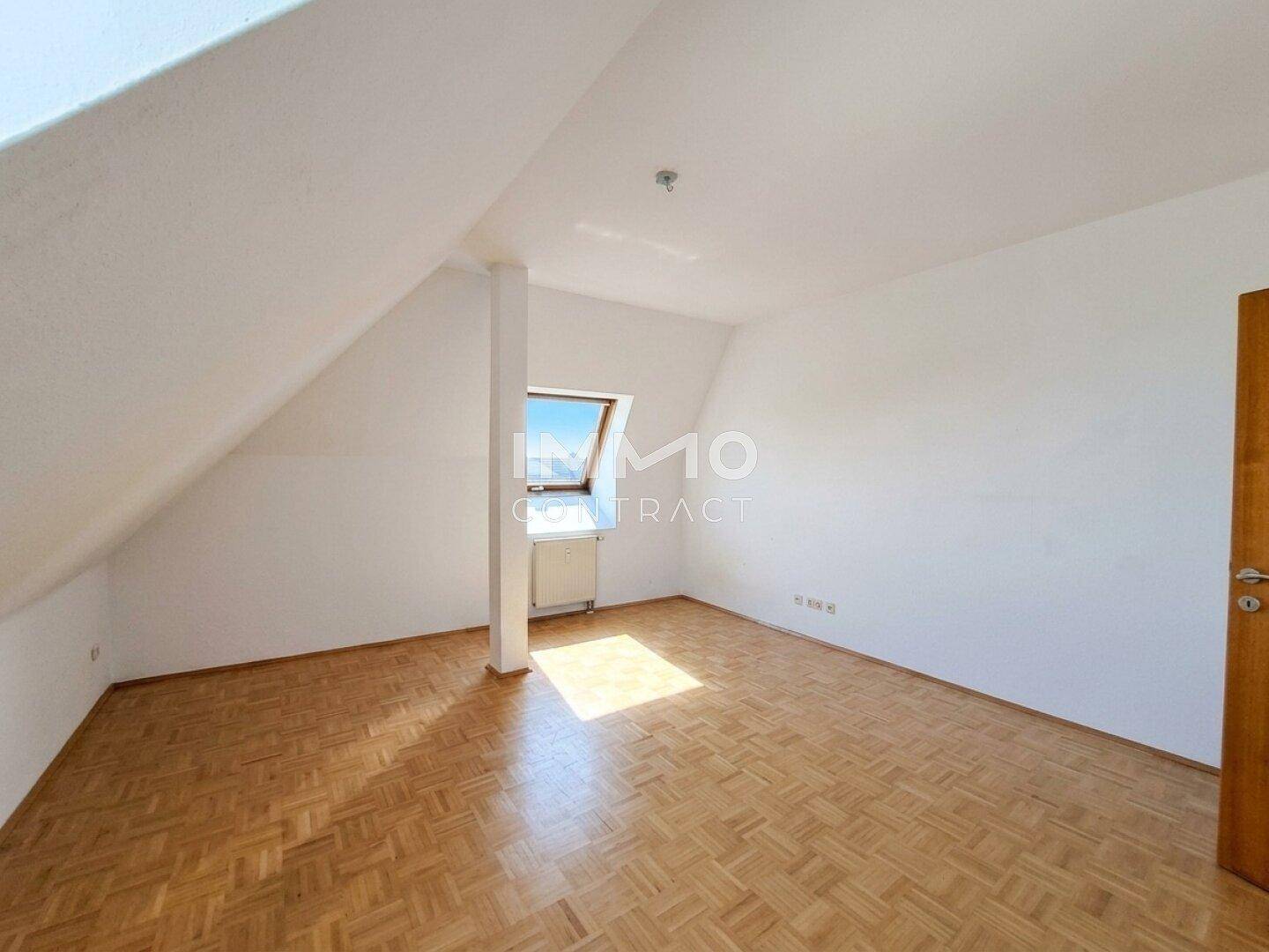 Helles Zimmer im Dachgeschoss mit schrägen Wänden, Parkettboden und einem Dachfenster.
